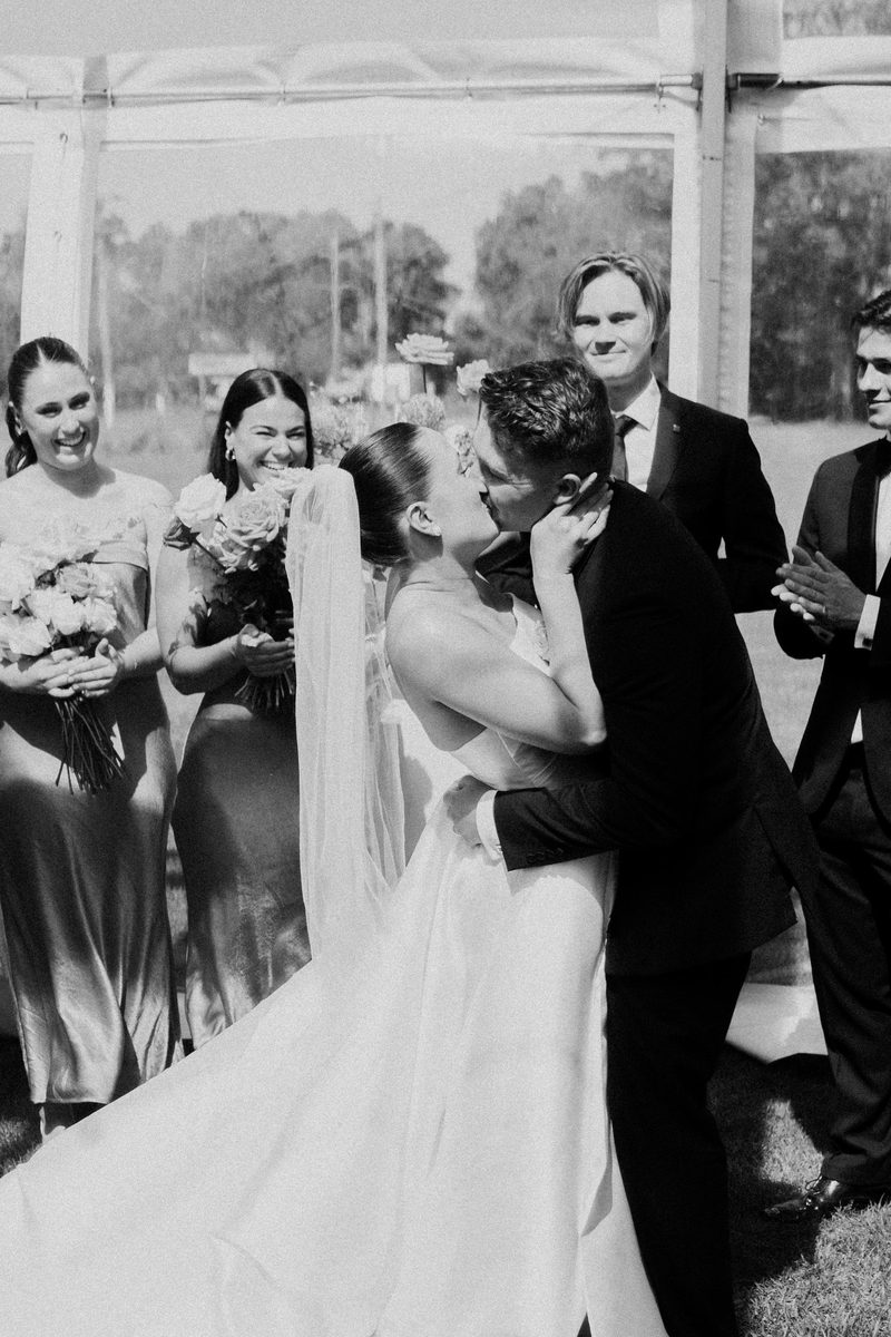 First kiss beside the bridesmaids, in black and white