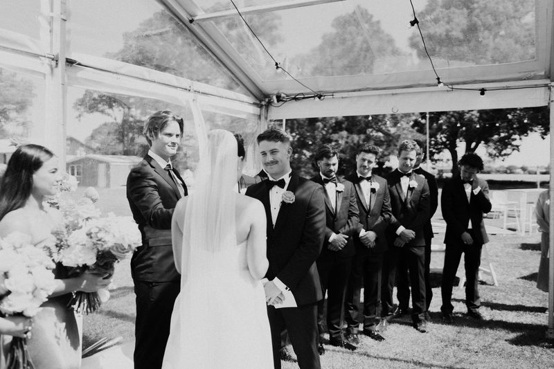 Groomsmen laughing with the couple in the marquee, in black and white